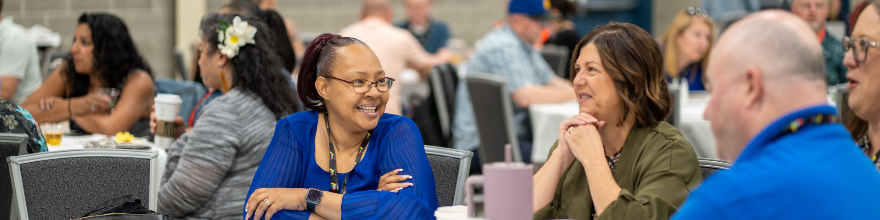 Connect Photo Only Webpage Header Groups of people at tables featuring a black woman in the center with a blue shirt smiling at others at the table.